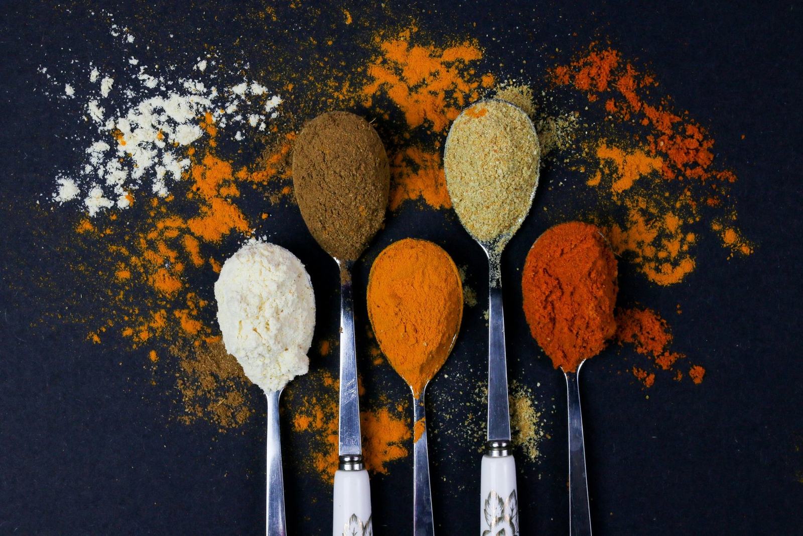Spoons of ground spices on a dark surface: turmeric, chilli, cumin, coriander and garlic powder